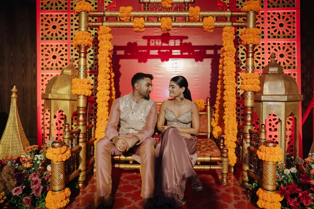Apeksha and Akash seated on the gold mehendi swing at Riva Beach Resort, Goa, framed by marigold strings, gold lanterns, red jaali screens and pink roses.