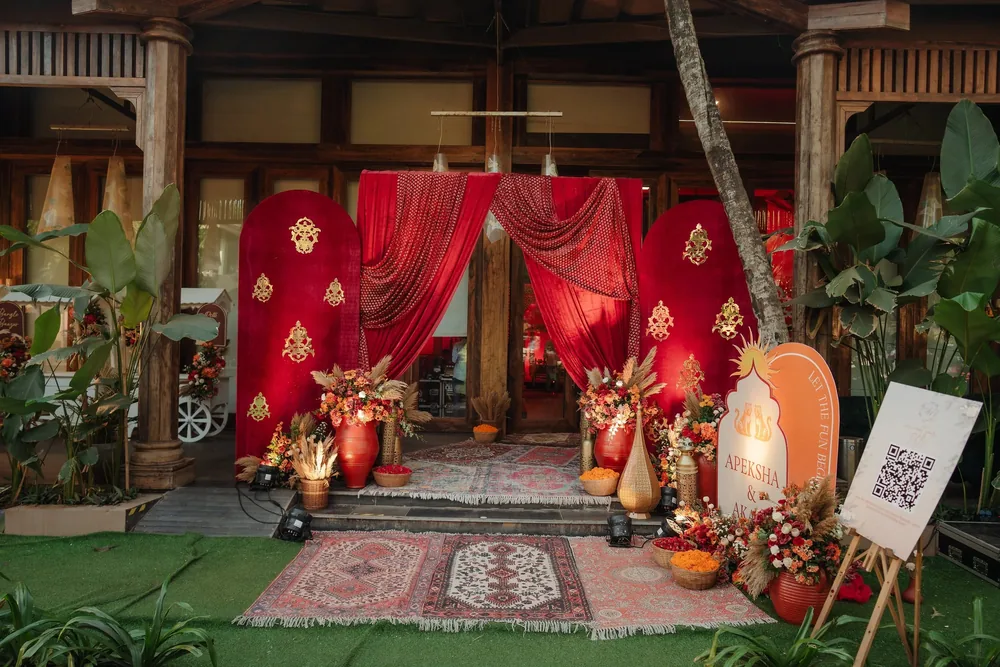 Mehendi entrance at Riva Beach Resort, Goa: swagged red velvet drapes, red arched panels with gold motifs, layered rugs and urns of orange blooms and pampas.