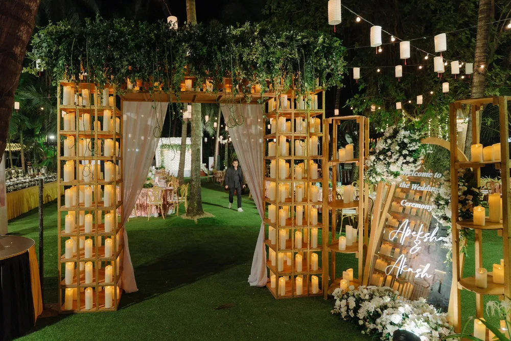 Reception entrance at Riva Beach Resort, Goa: wooden shelf towers of pillar candles, sheer blush drapes, greenery and white flowers under paper lanterns.