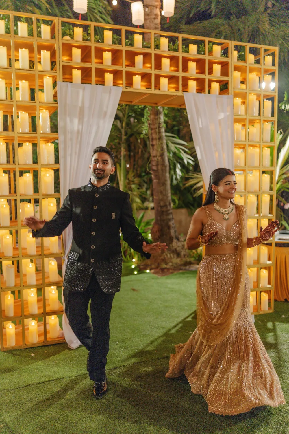 Apeksha and Akash walk into the reception through the candle-filled shelf gateway and sheer white drapes at Riva Beach Resort, Goa, under paper lanterns.