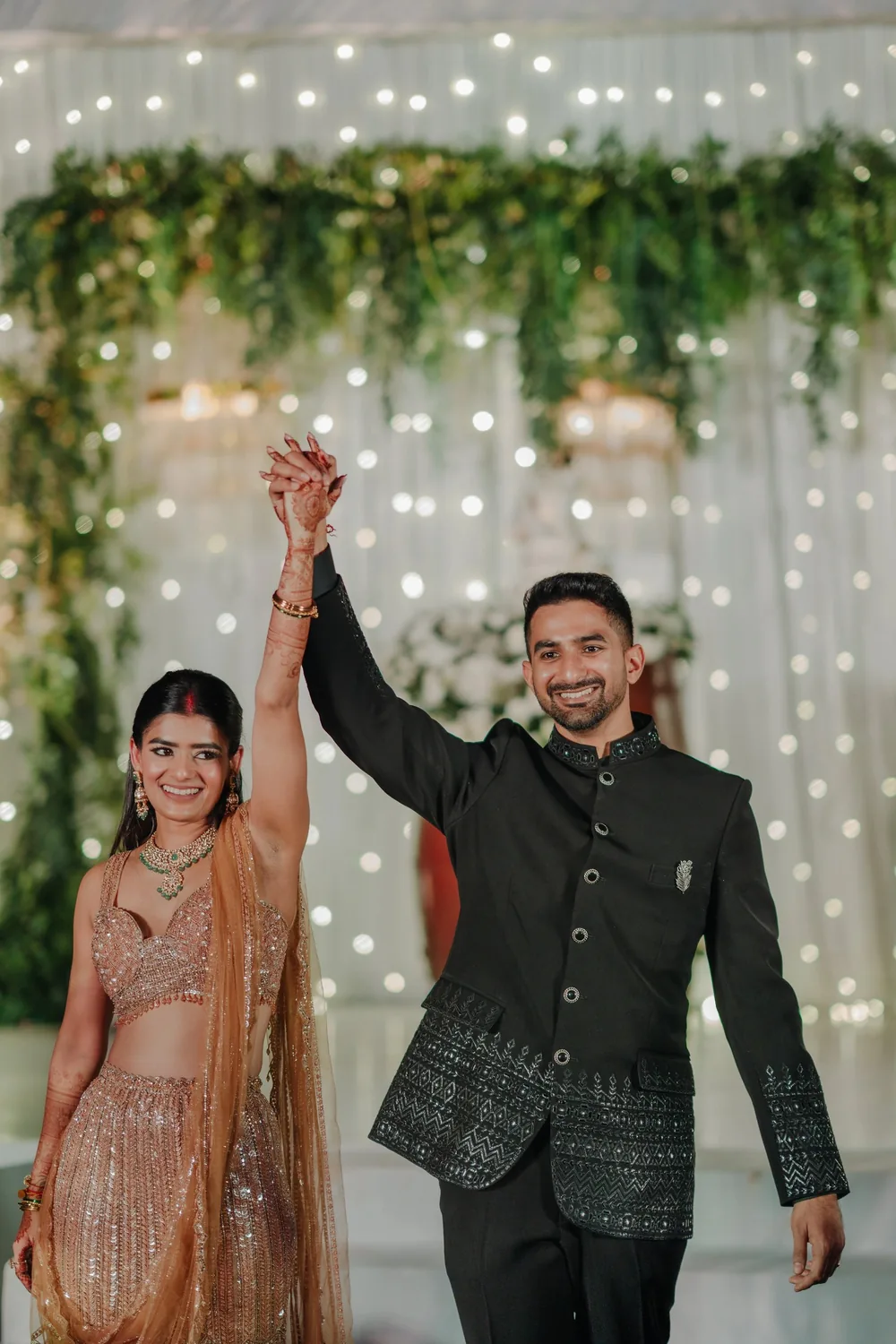 Apeksha and Akash raise joined hands on the reception stage at Riva Beach Resort, Goa, in front of a fairy-light curtain draped with trailing greenery.