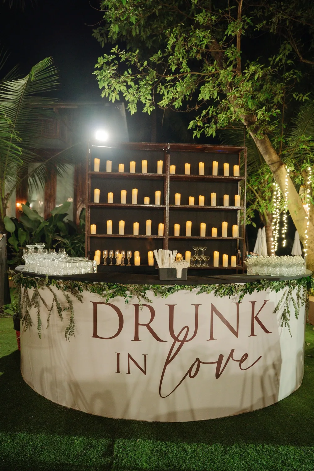 Reception bar at Riva Beach Resort, Goa: a round white counter lettered 'Drunk in Love' and trailed with greenery, backed by wooden shelves of pillar candles.