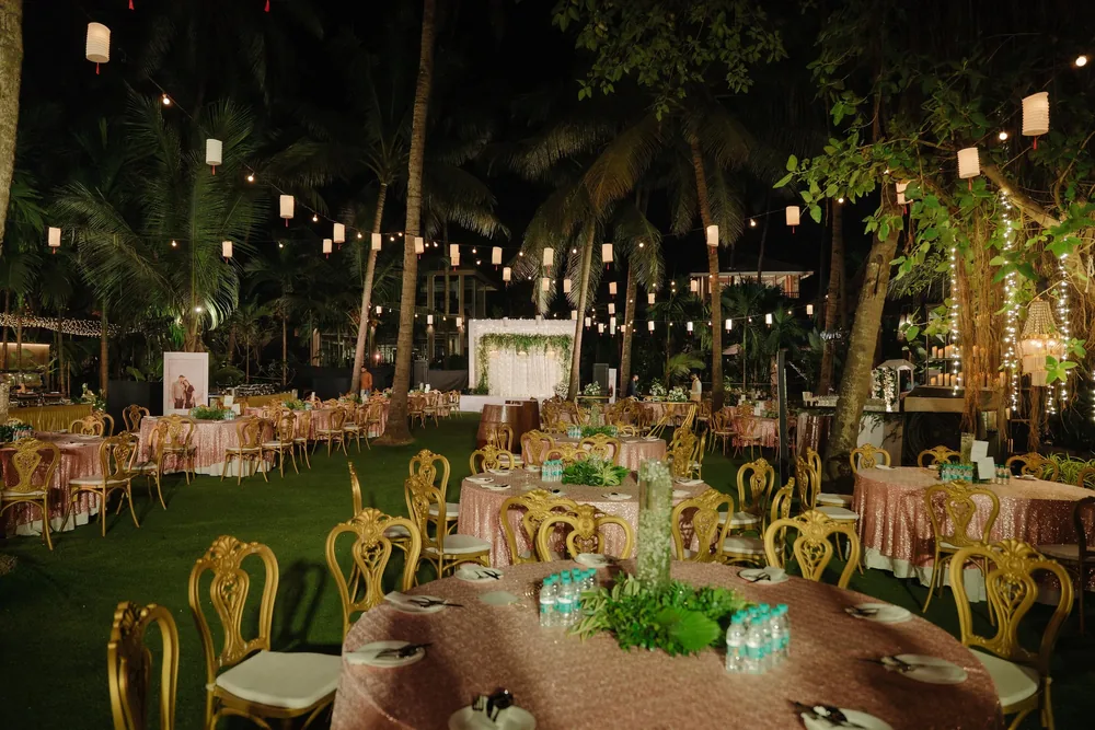 Reception lawn at Riva Beach Resort, Goa: round tables in rose-gold sequin cloths, gold carved chairs and greenery centrepieces under paper lanterns.