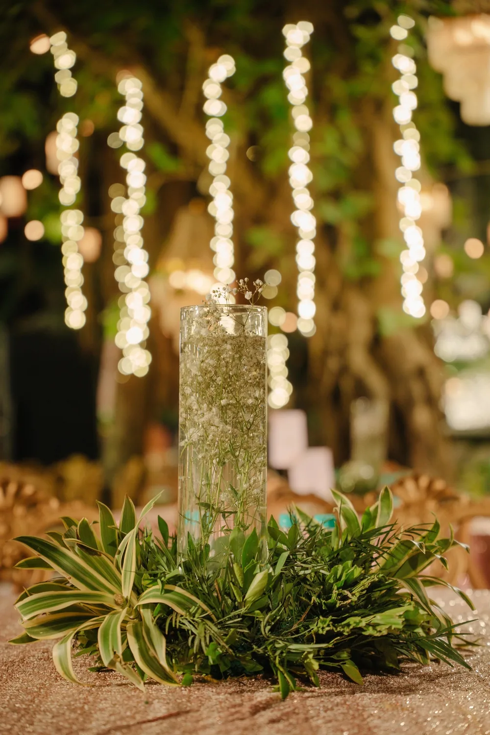 Reception centrepiece at Riva Beach Resort, Goa: a tall glass cylinder of baby's breath in a ring of leaves on rose-gold sequins, fairy lights behind.