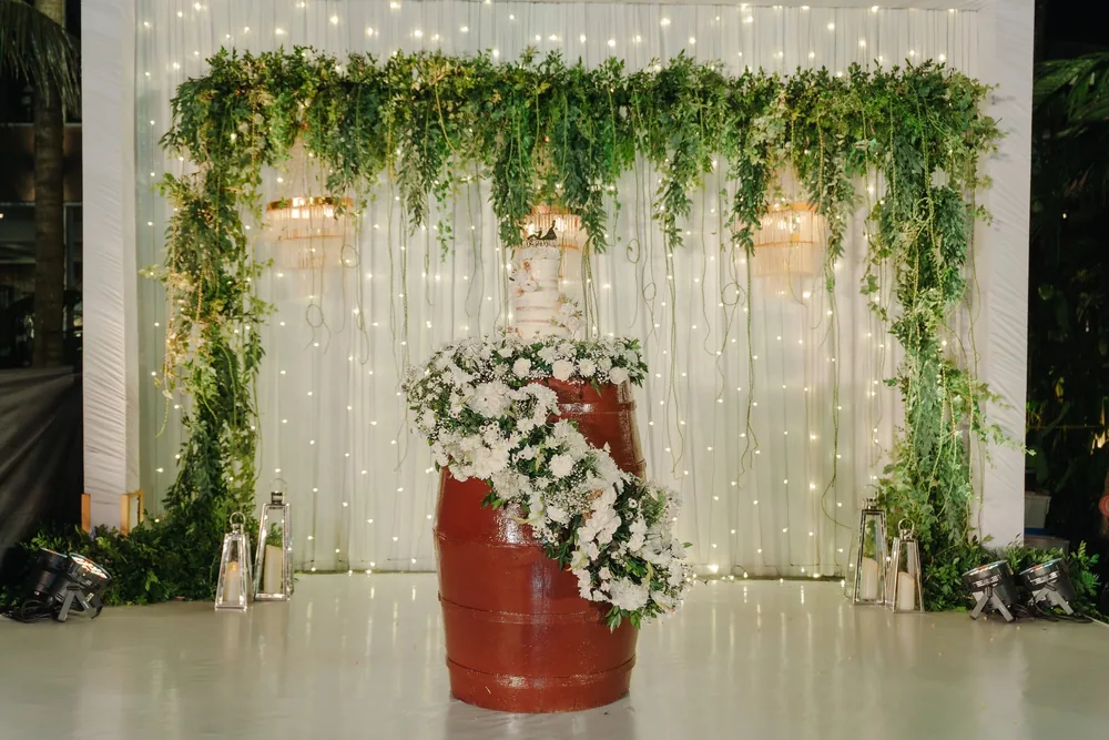 Reception stage at Riva Beach Resort, Goa: a fairy-light curtain with trailing greenery and chandeliers, and a red barrel spilling white flowers under the cake.