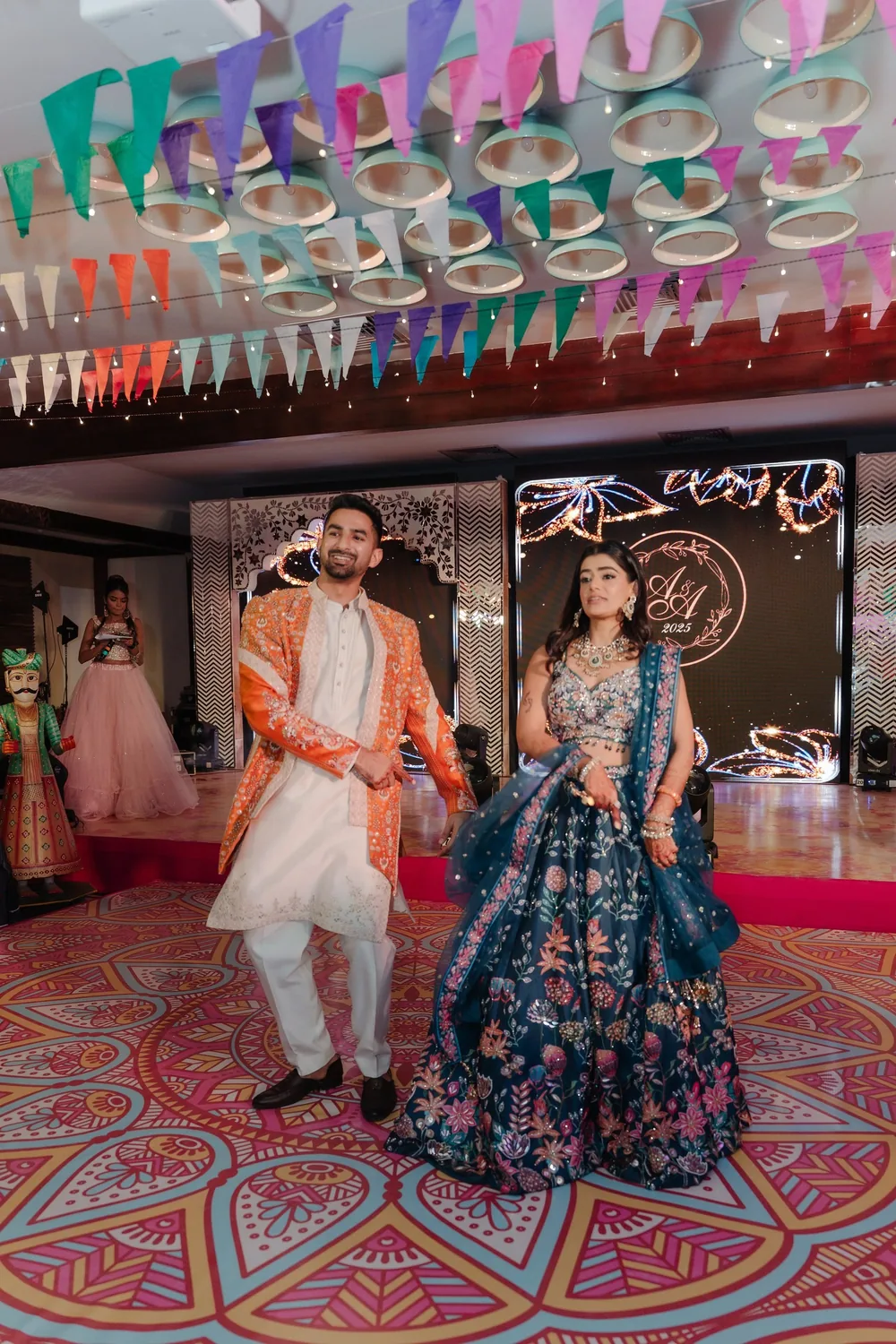 Apeksha and Akash dance on the mandala-print floor at the sangeet at Riva Beach Resort, Goa, under bunting, with the monogram screen and a puppet behind.
