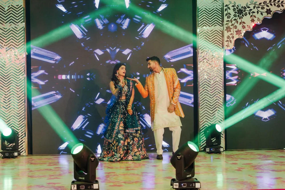 Apeksha and Akash perform on the sangeet stage at Riva Beach Resort, Goa, between white jaali panels, with LED screen visuals and green light beams.