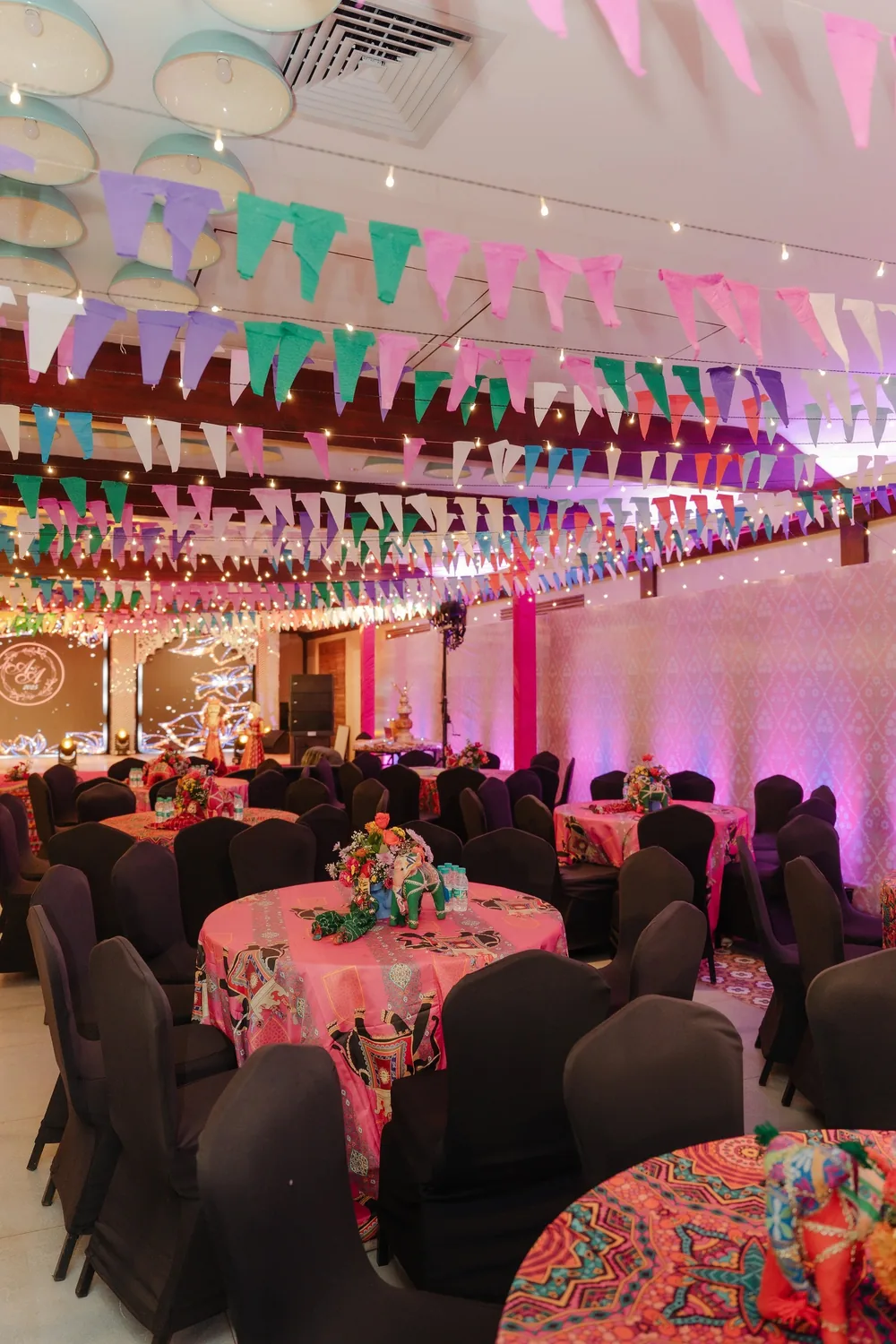 Sangeet hall at Riva Beach Resort, Goa: multicolour bunting and fairy lights above round tables in pink folk-print cloths with flowers and toy elephants.