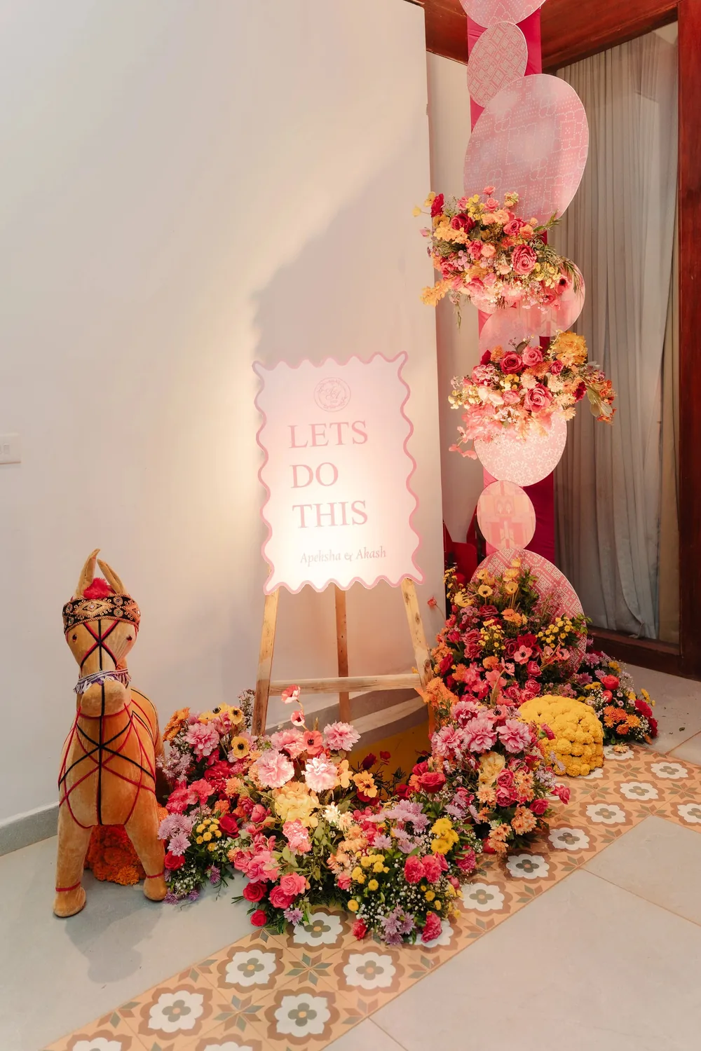 Sangeet sign at Riva Beach Resort, Goa: a pink scalloped 'Lets Do This' board on an easel beside pink printed discs, bright mixed flowers and a fabric camel.