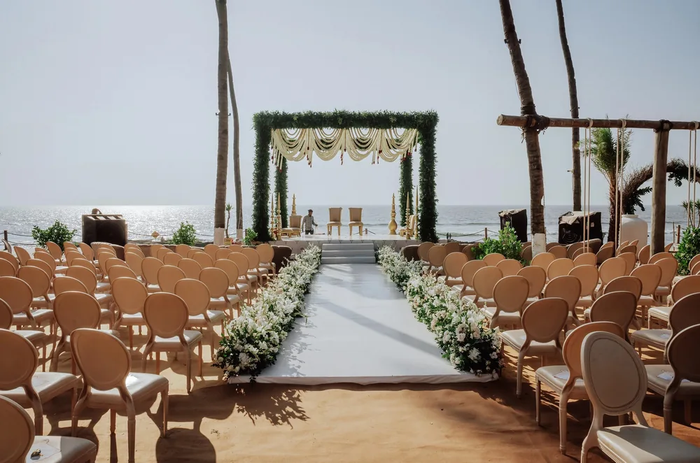 Wedding aisle at Riva Beach Resort, Goa: a white runner edged with white orchids, roses and greenery between cream chairs on the sand, leading to the mandap.