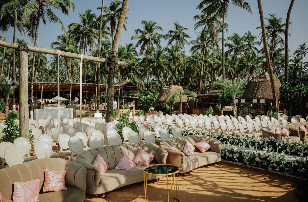 Wedding lounge at Riva Beach Resort, Goa: curved taupe velvet sofas with blush sequin cushions, a gold side table and rows of white chairs under coconut palms.