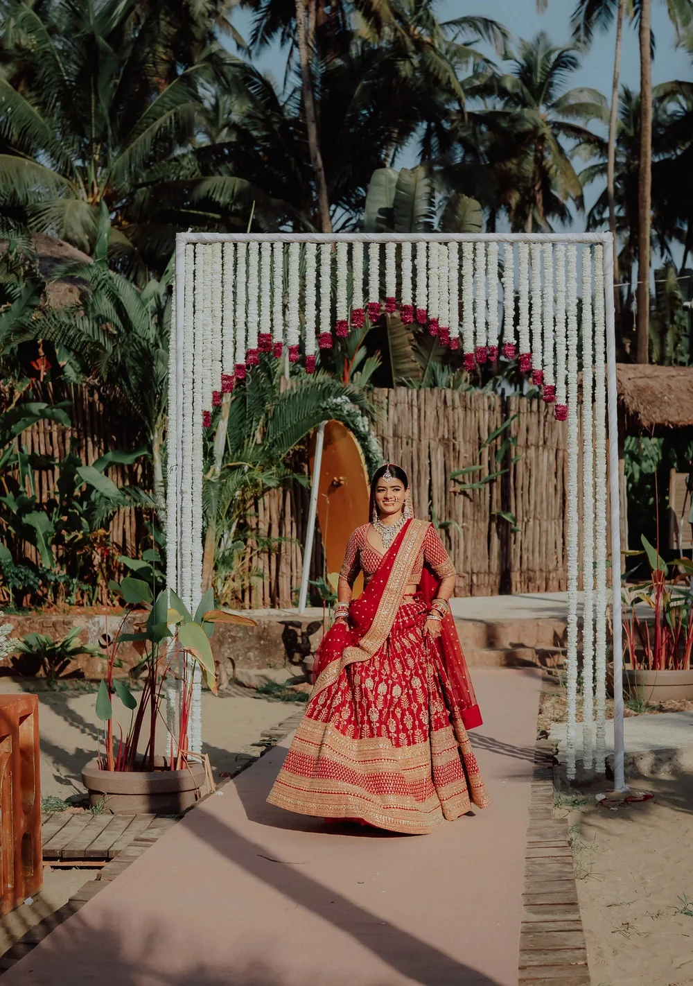 Apeksha in a red lehenga walks out under a white flower-string arch with an inner arc of red flowers on the wedding walkway at Riva Beach Resort, Goa.