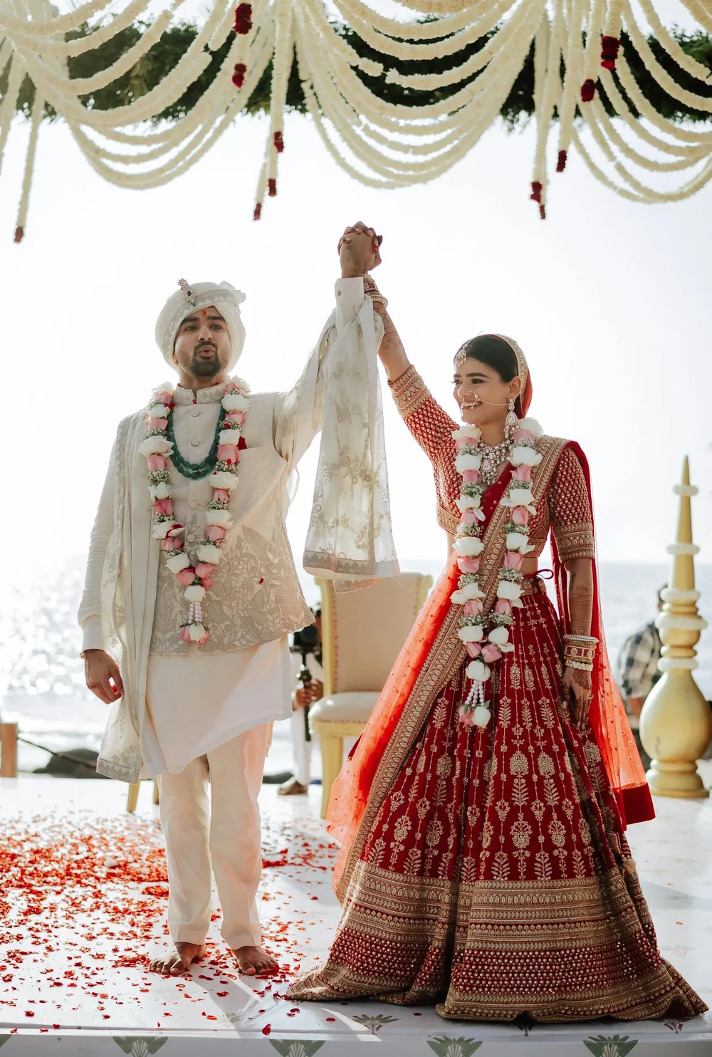 Apeksha and Akash raise joined hands under the wedding mandap swags at Riva Beach Resort, Goa, in rose garlands, with red petals across the white floor.
