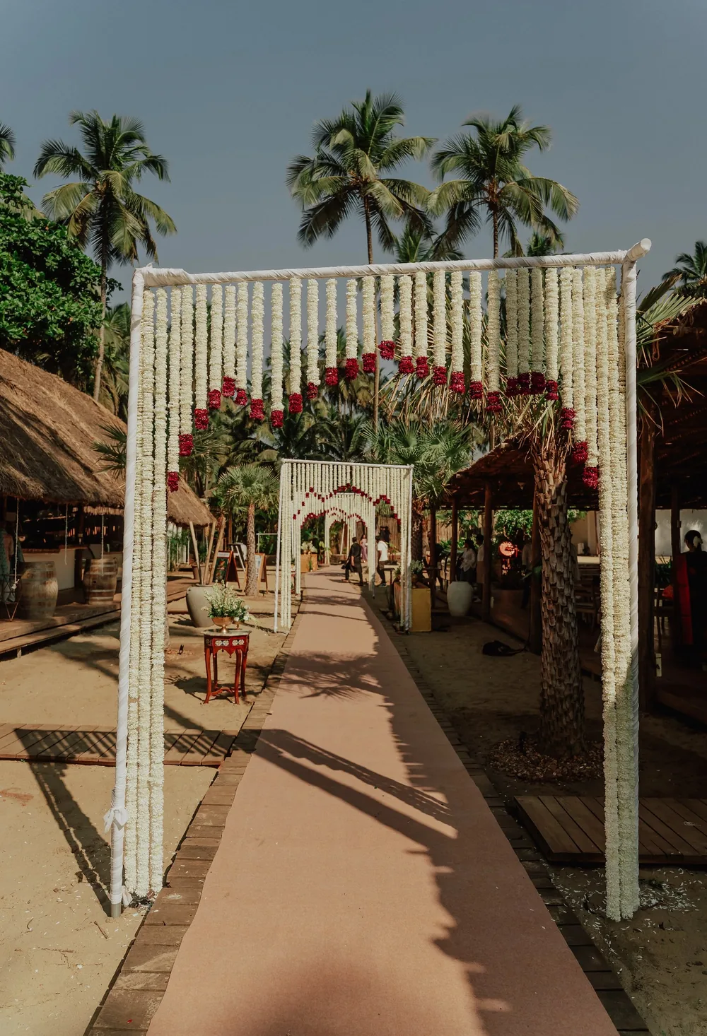 Wedding entrance at Riva Beach Resort, Goa: a run of white flower-string arches with inner arcs of red flowers over a walkway between palms and thatched huts.