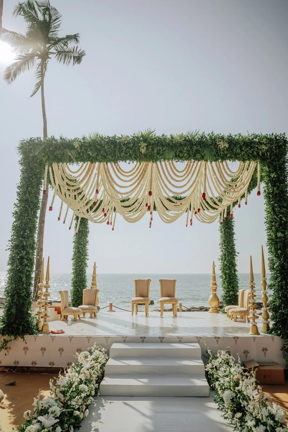 Wedding mandap at Riva Beach Resort, Goa: a foliage-covered frame hung with white flower-strand swags tipped in red, gold finials and cream chairs by the sea.