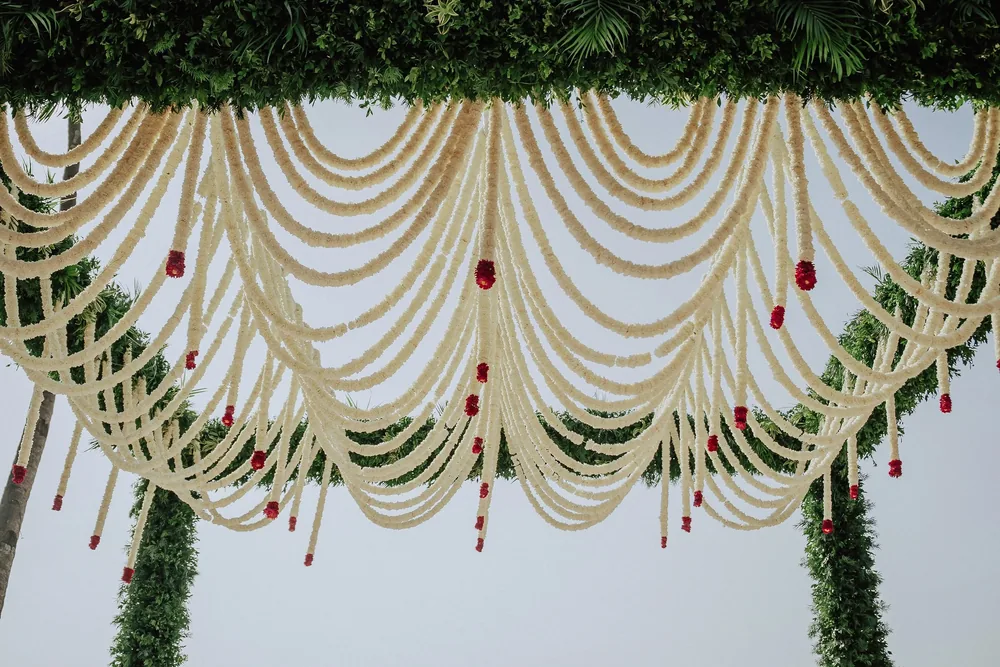 Wedding mandap detail at Riva Beach Resort, Goa: layered scallops of cream flower strands with red flower tassels, hung from a foliage frame against the sky.