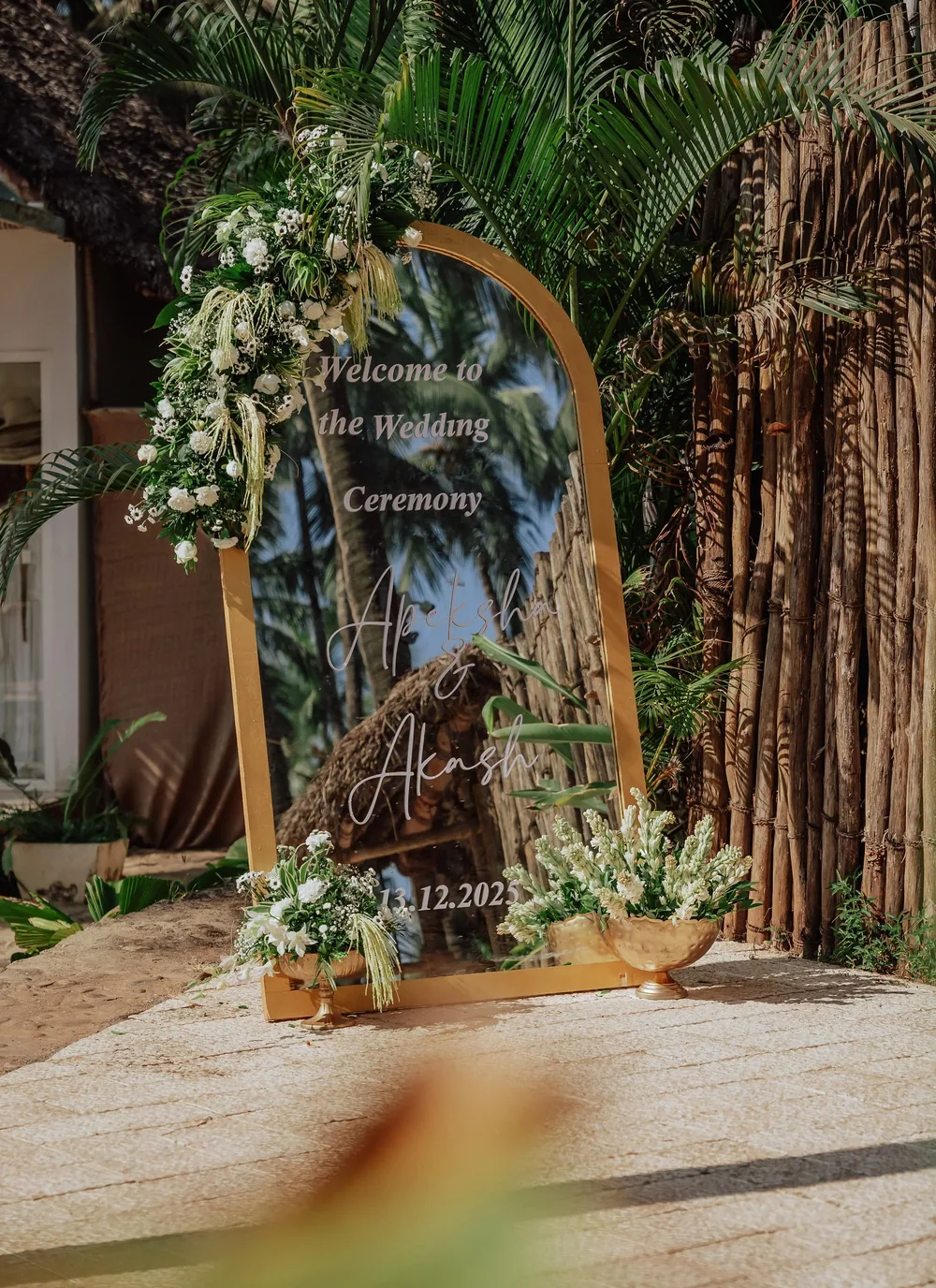 Wedding welcome sign at Riva Beach Resort, Goa: a gold-framed arched mirror with the couple's names, dressed in white roses, daisies and trailing amaranthus.