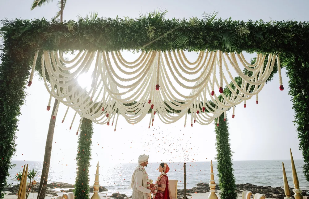 Rose petals fall over Apeksha and Akash under the wedding mandap's white floral swags at Riva Beach Resort, Goa, with gold finials and the sea beyond.