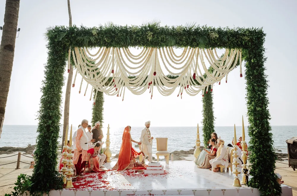 The wedding pheras under the foliage-and-swag mandap at Riva Beach Resort, Goa, as guests look on, with rose petals on the white floor and the sea behind.