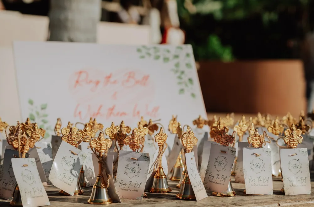 Wedding favours at Riva Beach Resort, Goa: brass bells tied with jute twine and A&A monogram tags reading 'Ring the Bell', in front of a matching sign.