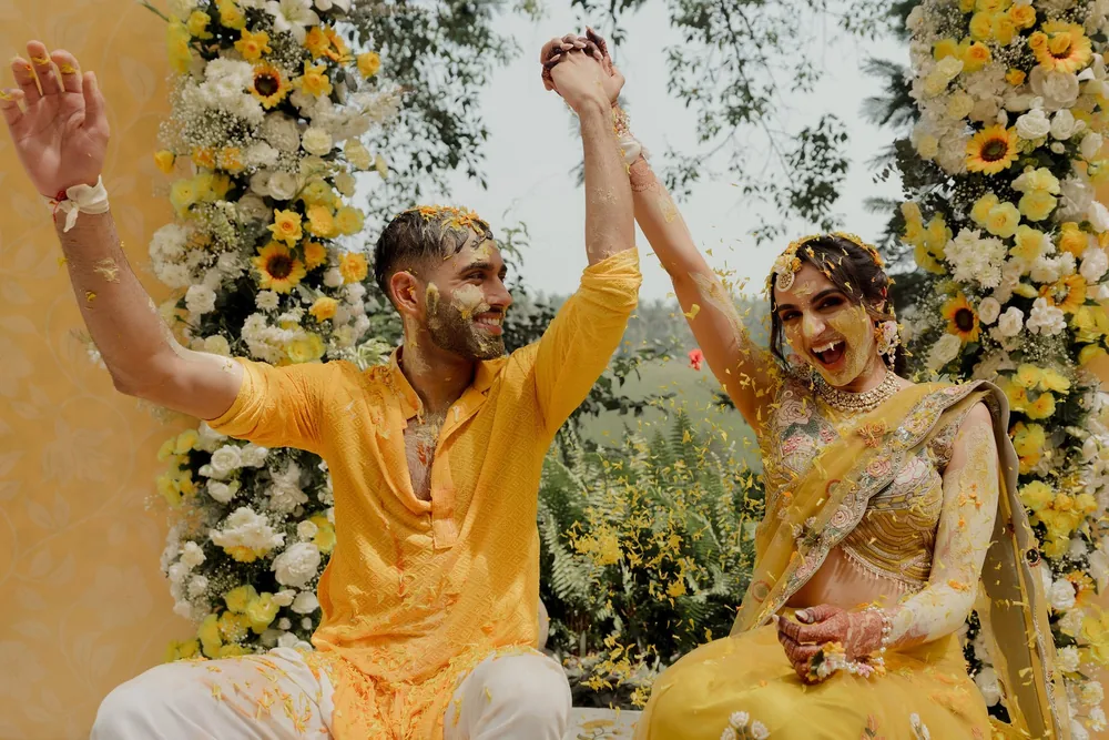 Shivani and Rohan, faces dabbed with haldi, raise joined hands between columns of sunflowers and white flowers at the haldi at Beleza by the Beach, Goa.