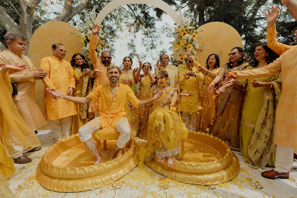 Family in yellow shower petals on Shivani and Rohan in gold tubs before the mustard arches and sunflower columns of the haldi at Beleza by the Beach, Goa.