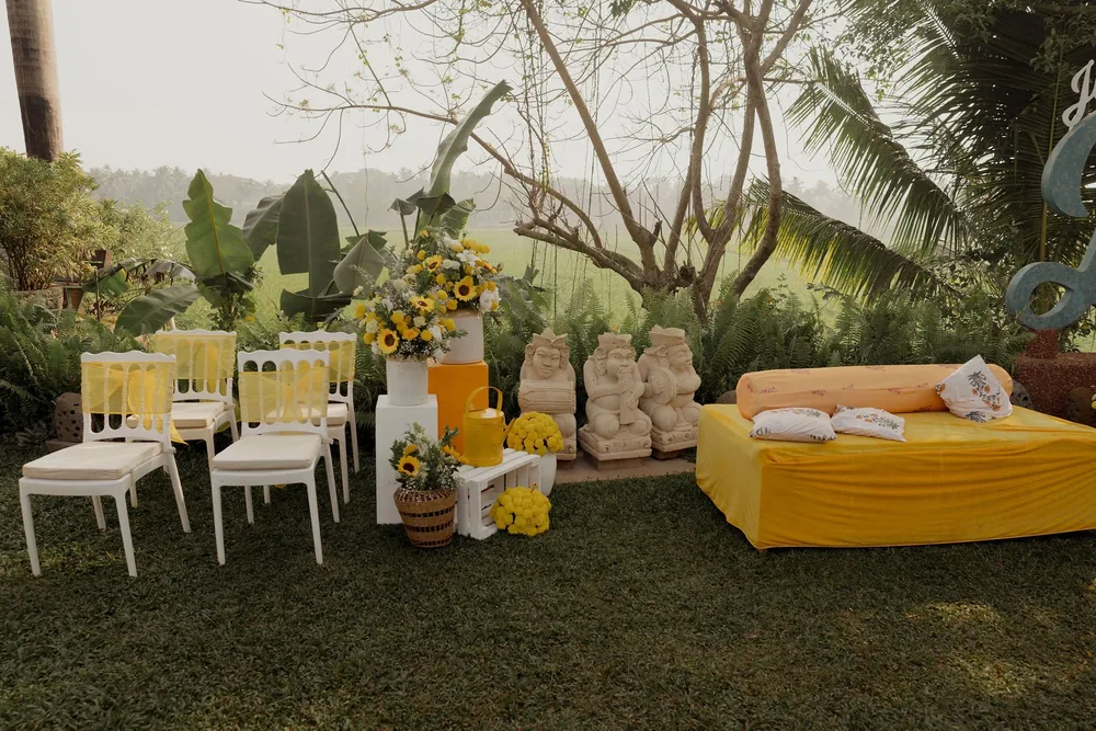 Haldi lounge at Beleza by the Beach, Goa: chairs with yellow organza sashes, sunflower pots, a watering can, stone statues and a yellow daybed.