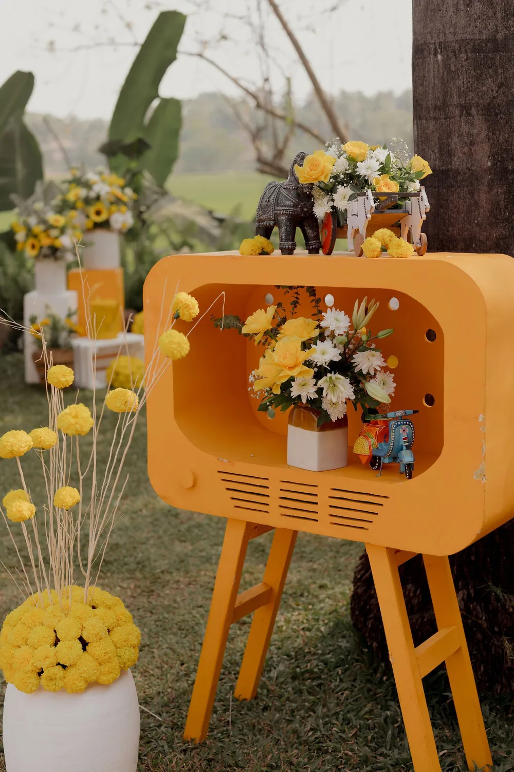 Orange retro TV-shaped stand at the haldi at Beleza by the Beach, Goa, with yellow roses, white blooms, a metal elephant, a toy cart and a painted toy scooter.
