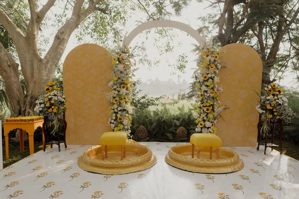 Haldi stage at Beleza by the Beach, Goa: mustard arched panels, a white ring arch, sunflower and white flower columns and two gold tubs on a printed floor.