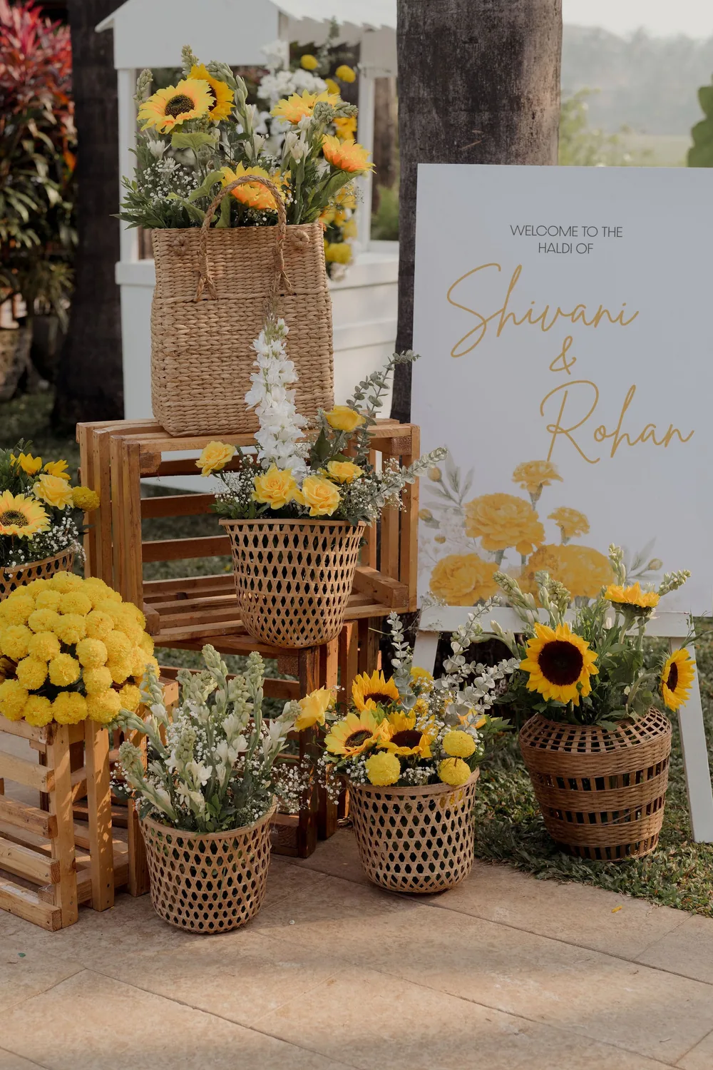 Haldi welcome sign for Shivani & Rohan at Beleza by the Beach, Goa, beside stacked wooden crates and wicker baskets filled with sunflowers and white flowers.