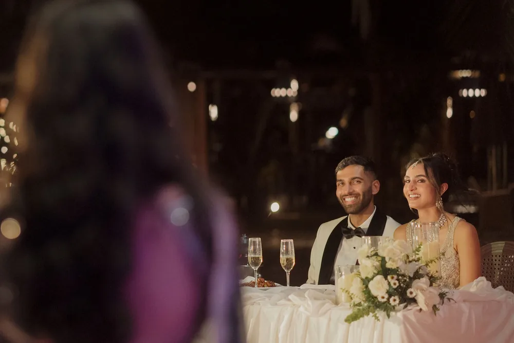 Shivani and Rohan at a white-draped sweetheart table with white roses, small daisies and candles in glass at the reception at Beleza by the Beach, Goa.