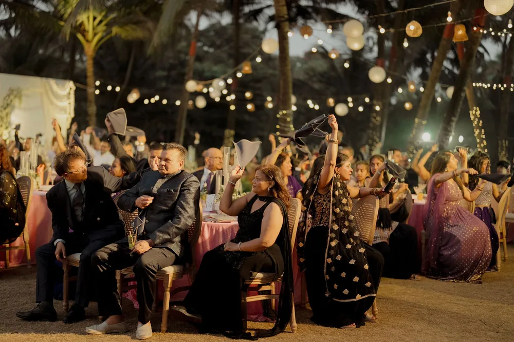 Reception guests wave napkins overhead at pink-clothed tables under festoon bulbs and paper lanterns strung through the palms at Beleza by the Beach, Goa.