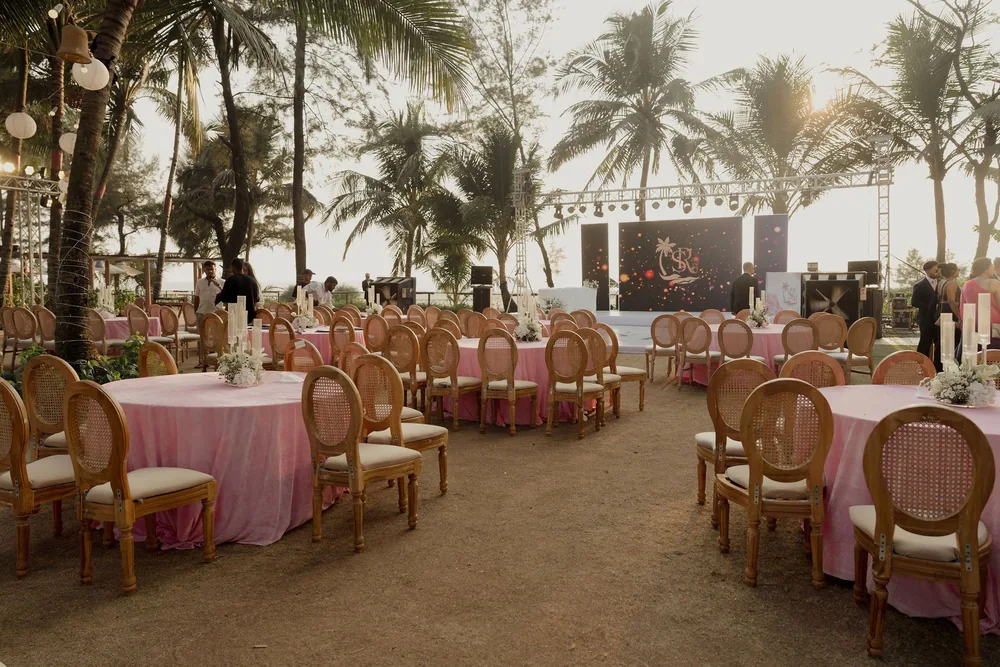 Reception lawn at Beleza by the Beach, Goa: round tables in pink cloths with cane-back chairs and glass candle centrepieces under palms at golden hour.