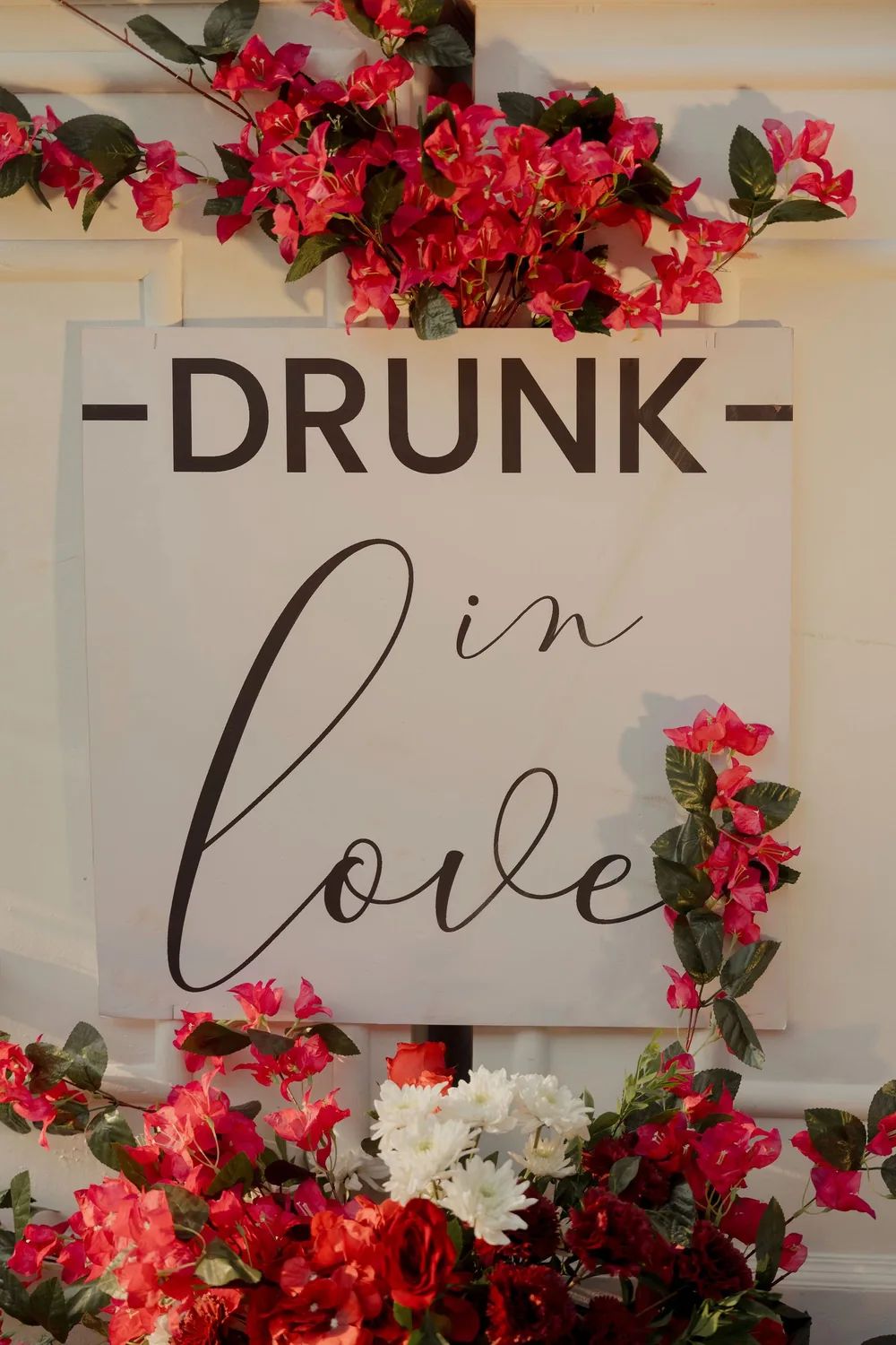 Bar sign reading Drunk in love at the sangeet at Beleza by the Beach, Goa, framed with magenta bougainvillea, red roses and white flowers on a white bar front.