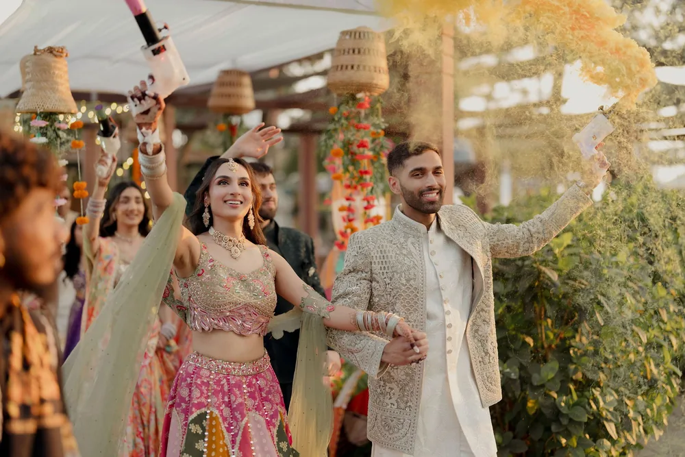 Shivani and Rohan enter the sangeet at Beleza by the Beach, Goa with colour smoke, under woven lampshades and hanging strings of orange and red flowers.