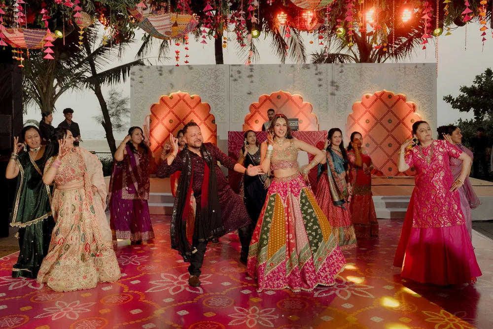 Shivani dances with family on a pink printed dance floor before the white stage, coral arches and umbrella canopy of the sangeet at Beleza by the Beach, Goa.