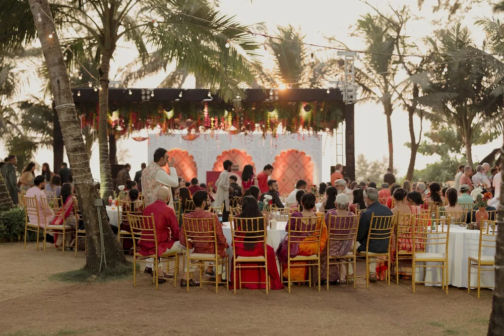 Guests at white-linen tables with gold chairs face the sangeet stage and its flower canopy under string lights and palms at Beleza by the Beach, Goa.