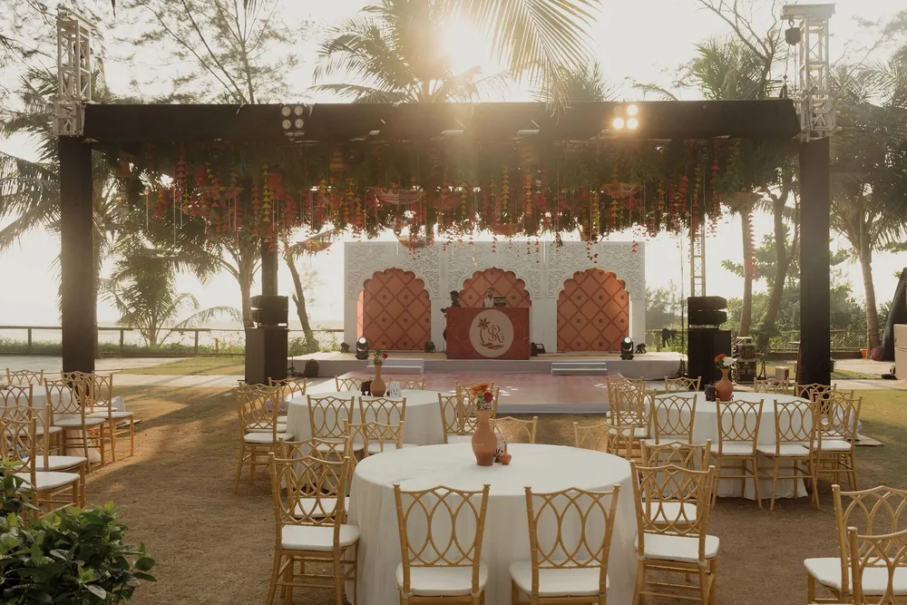 Sangeet lawn at Beleza by the Beach, Goa at golden hour: white-linen tables and gold chairs facing a carved white arched stage under a hanging canopy.