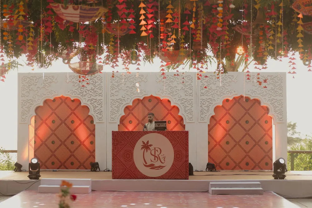 Sangeet stage at Beleza by the Beach, Goa: carved white panels with three scalloped coral arches, a monogrammed console and a canopy of umbrellas and tassels.