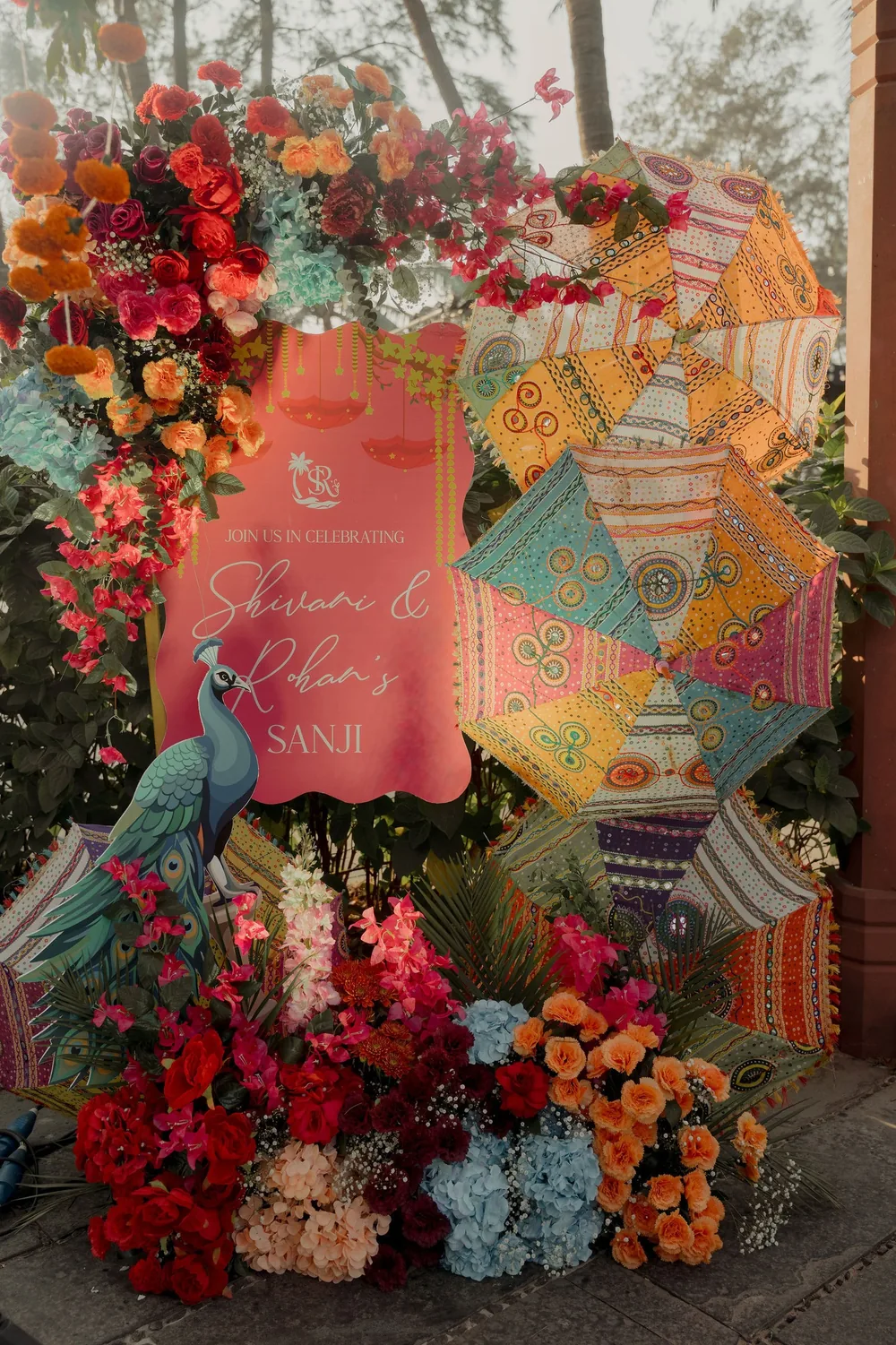 Coral sangeet sign for Shivani & Rohan's Sanji at Beleza by the Beach, Goa, with a peacock cut-out, embroidered umbrellas and red, orange and blue flowers.