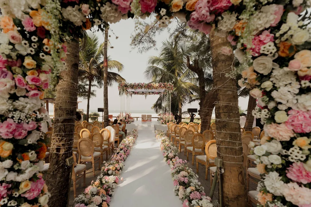 Through a floral arch of pink, peach and white roses, a flower-lined white aisle and cane-back chairs lead to the wedding mandap at Beleza by the Beach, Goa.