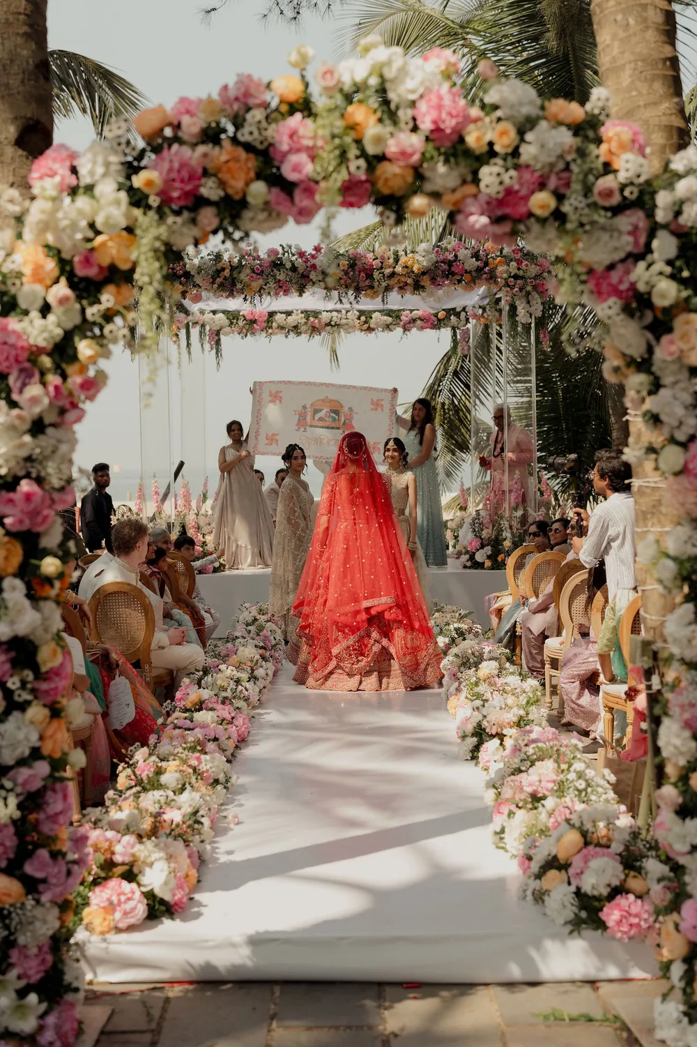 Shivani, in a red lehenga and veil, walks under a floral arch down the flower-lined aisle to the wedding mandap at Beleza by the Beach, Goa.