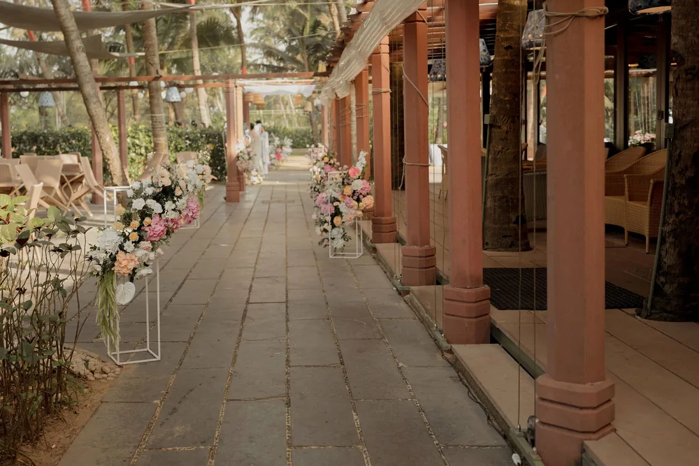 Stone walkway beside a red colonnade at Beleza by the Beach, Goa, lined for the wedding with white stands of pink, peach and white roses and hydrangeas.