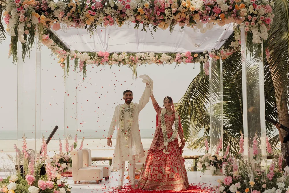 Shivani and Rohan raise joined hands as petals fall under the acrylic wedding mandap and its pink, peach and white floral canopy at Beleza by the Beach, Goa.