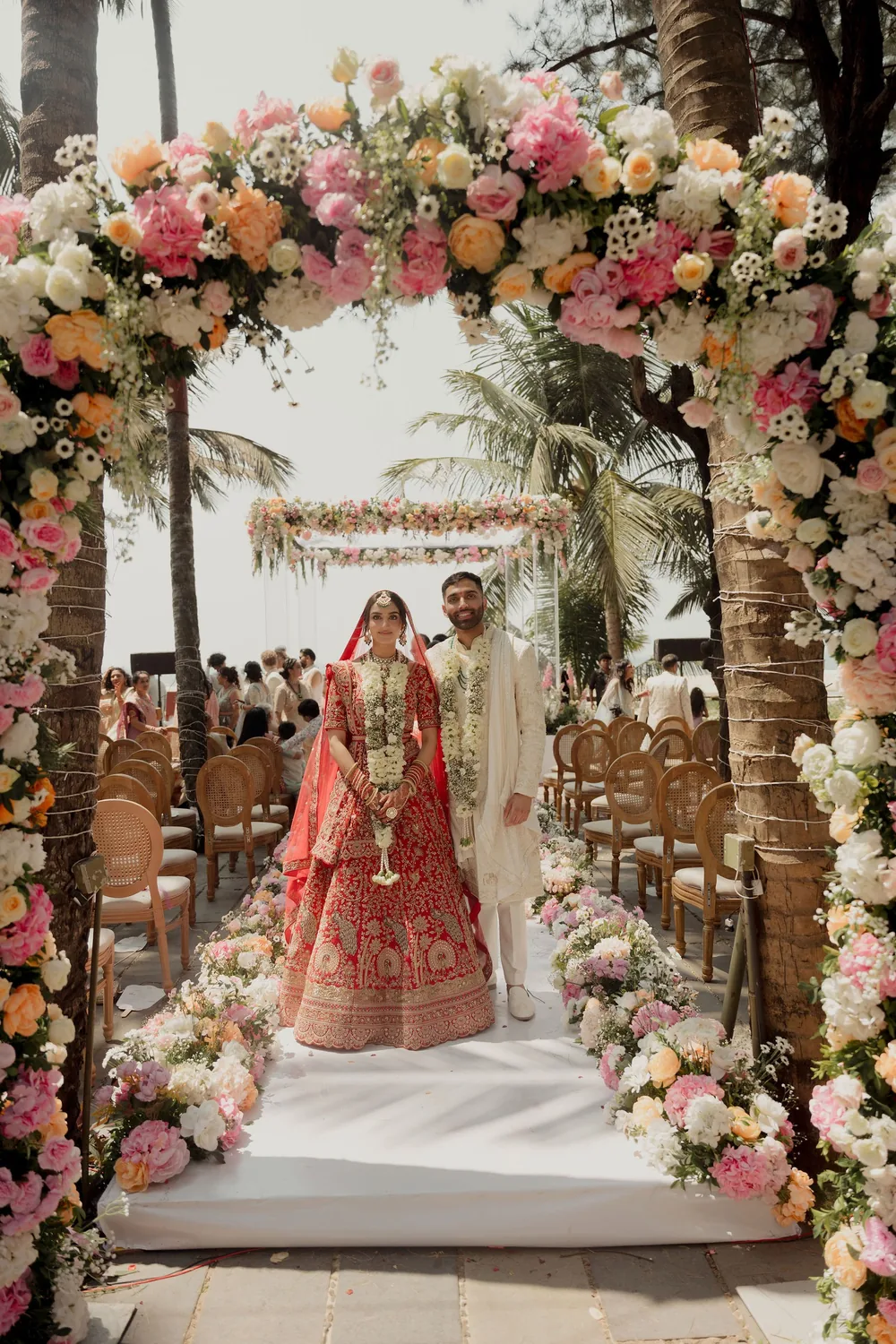 Shivani and Rohan stand under the pink, peach and white floral arch at the head of the wedding aisle, the mandap behind them, at Beleza by the Beach, Goa.
