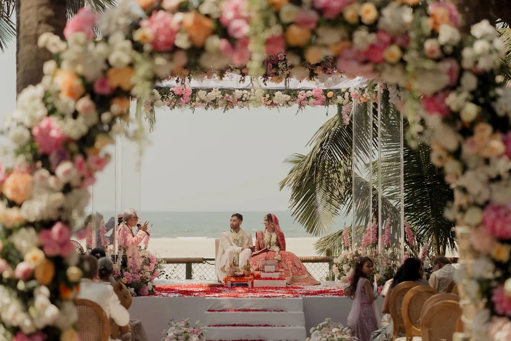 Floral arch framing the wedding mandap at Beleza by the Beach, Goa, where Shivani and Rohan sit on a petal-strewn white platform with the sea behind them.