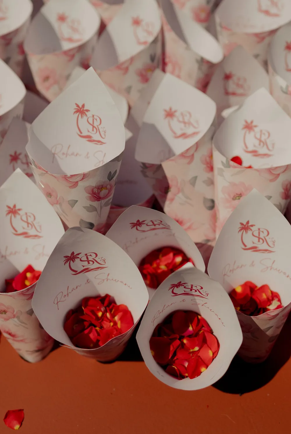 Paper cones printed with the couple's palm-tree monogram and pink florals, filled with red rose petals for the wedding ceremony at Beleza by the Beach, Goa.