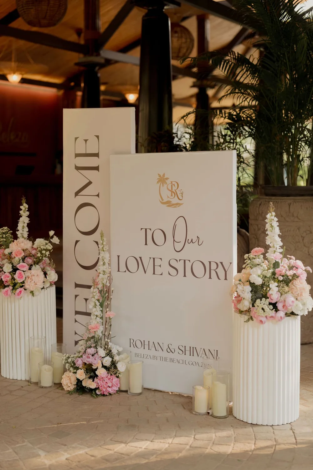 Wedding welcome boards for Rohan & Shivani at Beleza by the Beach, Goa, between fluted pedestals of pink and white roses, with pillar candles in glass.