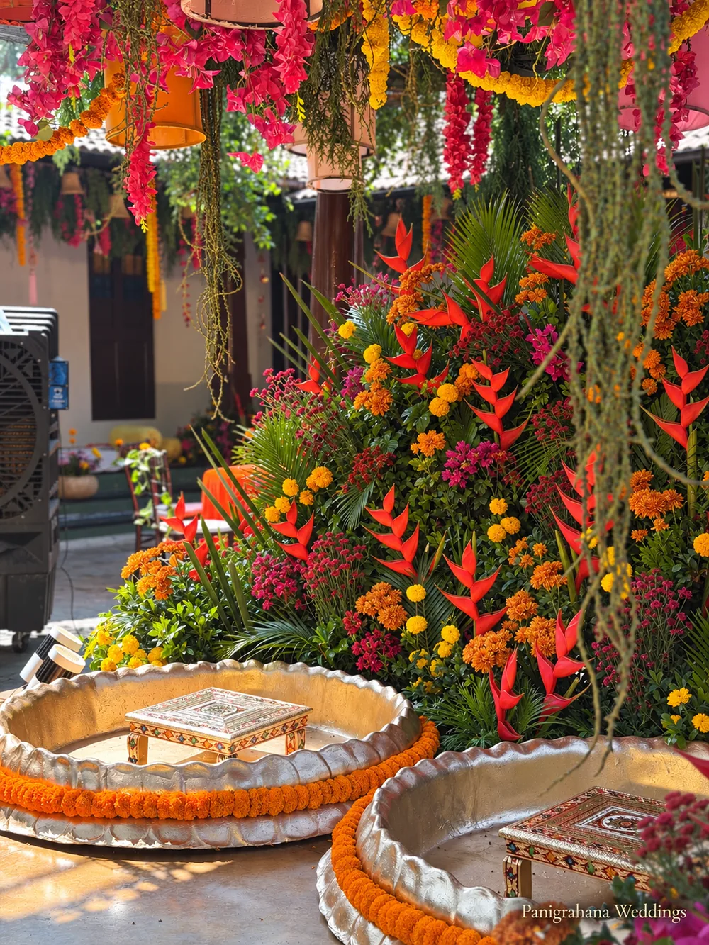 Close view of the haldi stage: brass urlis with marigold rims and small wooden stools, red heliconia, hanging amaranthus and orange and pink flowers