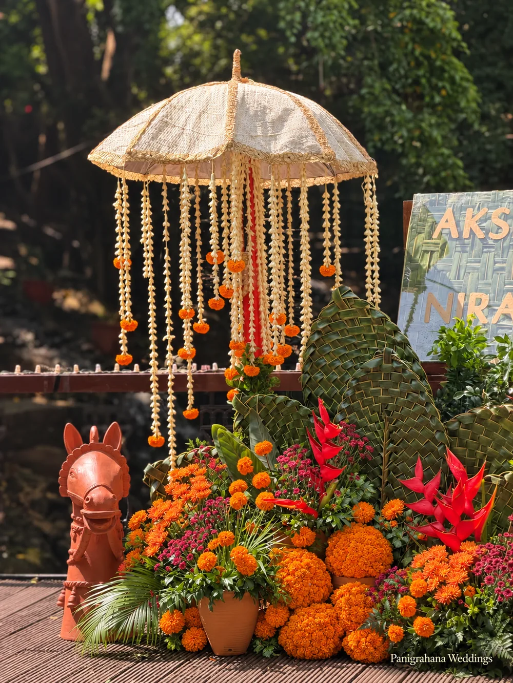 A cream chhatri hung with marigold and jasmine strings above woven coconut-leaf mounds, a terracotta horse, red heliconia and marigold heaps at a haldi in Bangalore